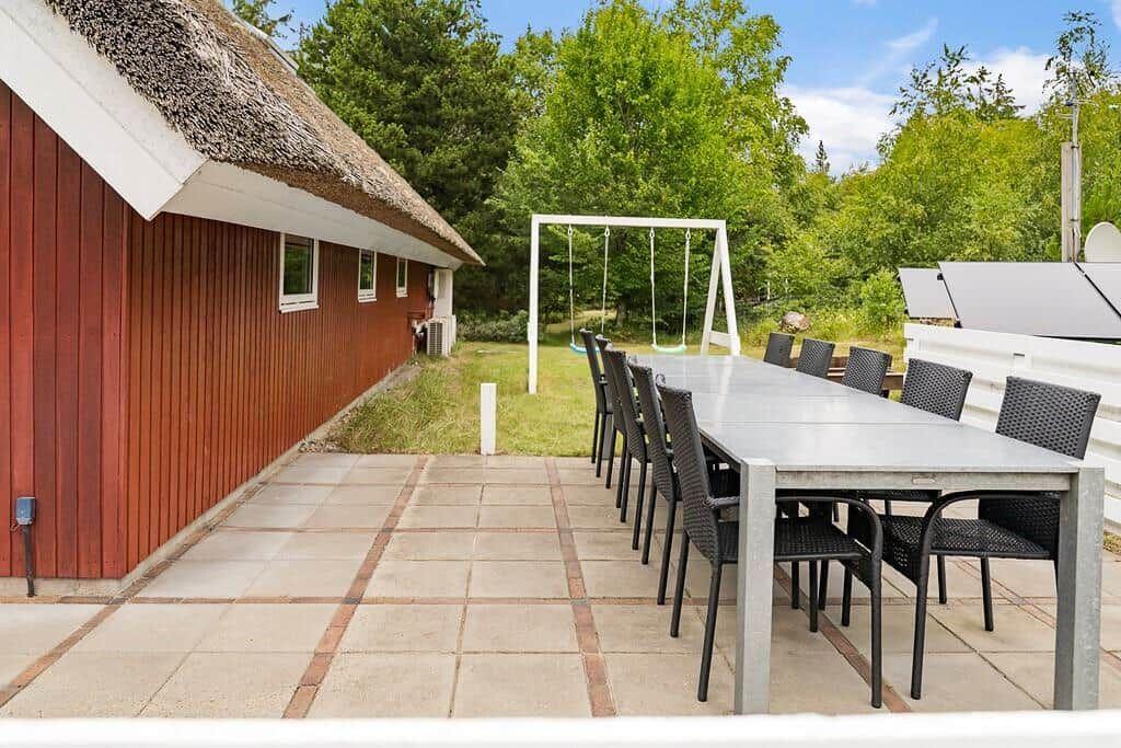 Terrasse mit langem Tisch und Stühlen, daneben Haus mit Strohdach und Schaukel im Hintergrund.