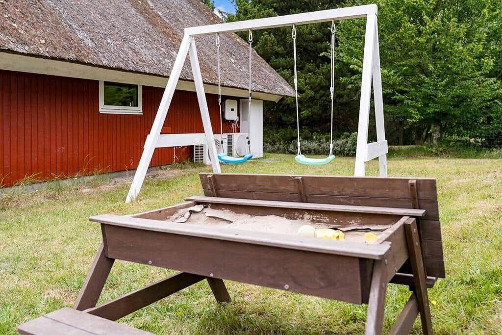 Hinter dem roten Haus steht ein weißer Schaukelstand mit zwei Schaukeln und ein Holzspielplatz mit Sand.