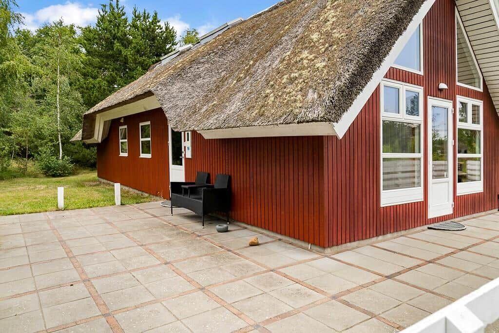 Rotes Holzhaus mit Strohdach und Terrasse. Schwarze Sitzgelegenheit vor dem Eingang.