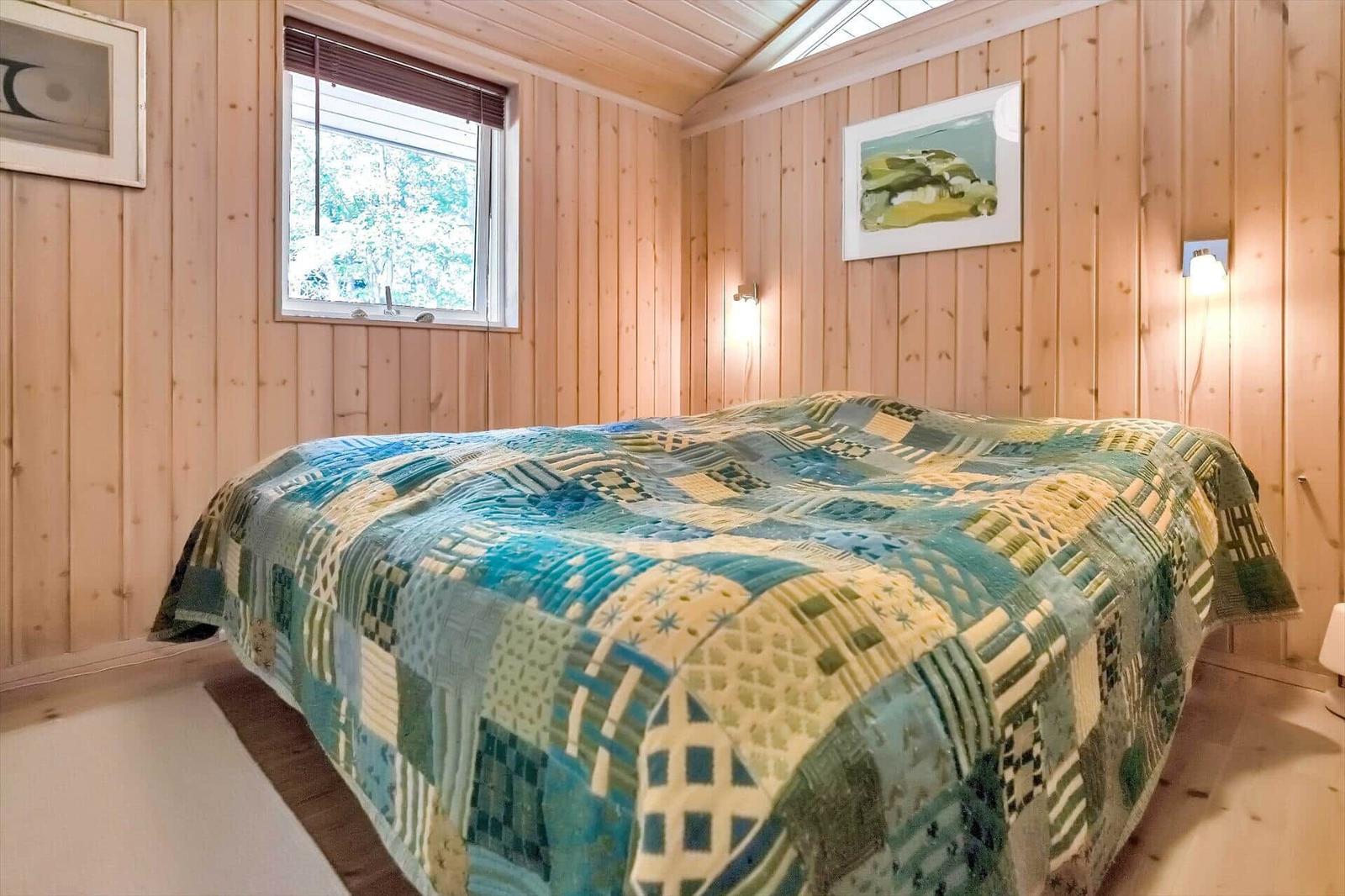 Schlafzimmer mit Holzwand, Bett mit Patchwork-Bettwäsche und Fenster mit Blick auf Bäume.