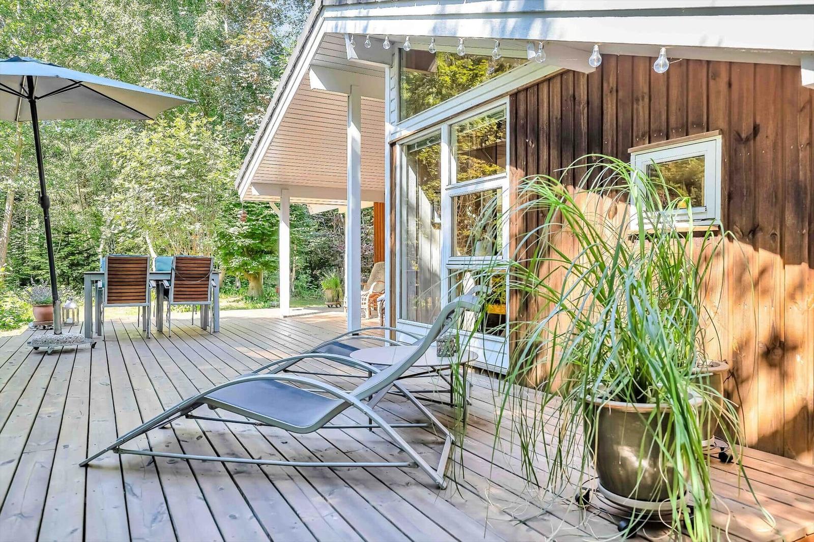 Terrasse mit Liegestuhl, Tisch, Stühlen und Pflanze vor Holzhäusern.