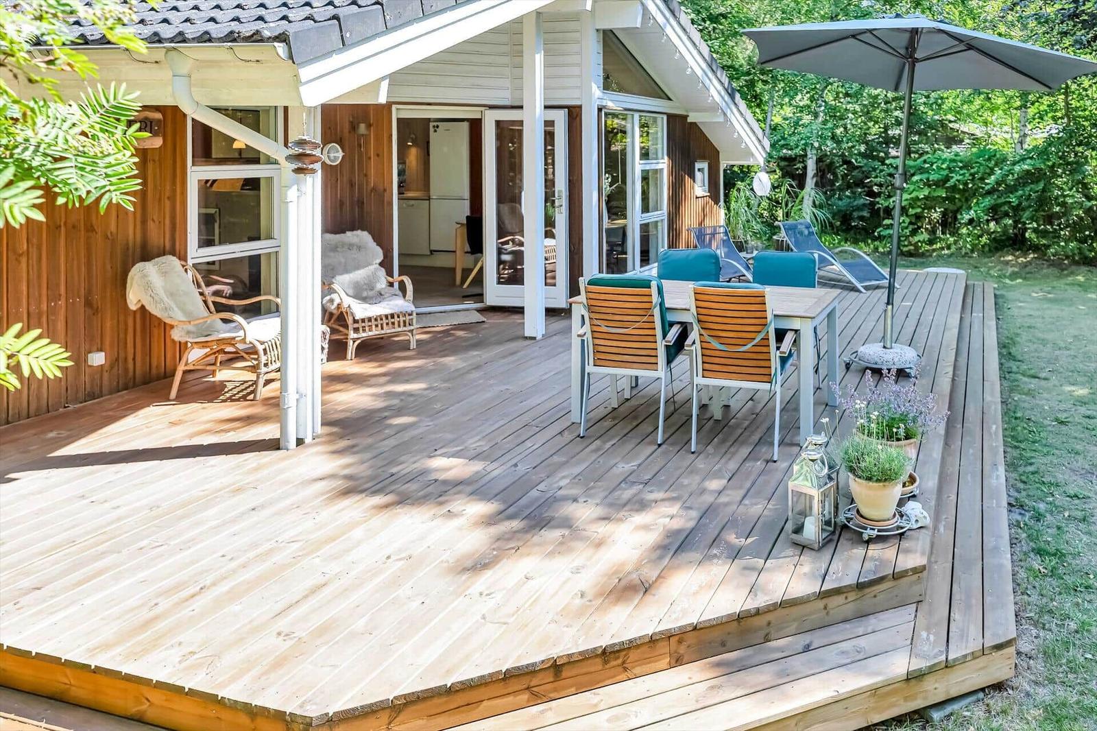 Holzterrasse mit Sitzgruppe, Sonnenschirm und Pflanzen. Blick in das Haus mit Küchenbereich.