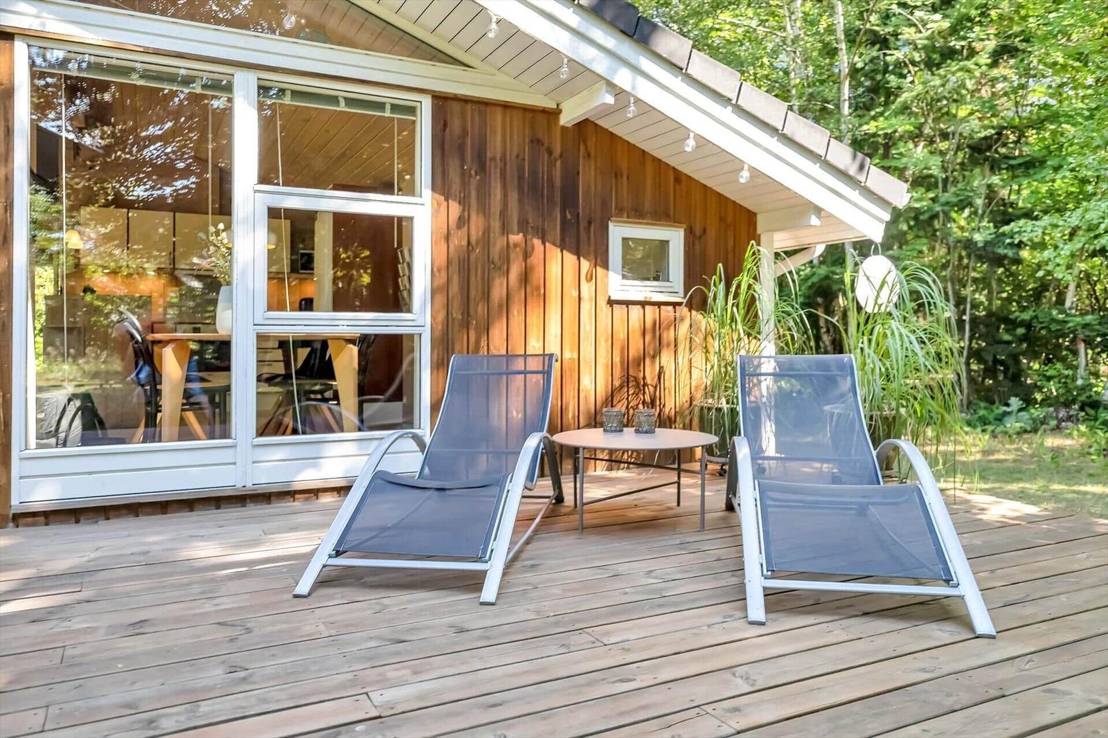 Terrasse mit zwei Liegestühlen und Tisch vor Holzhaus mit großem Fenster.