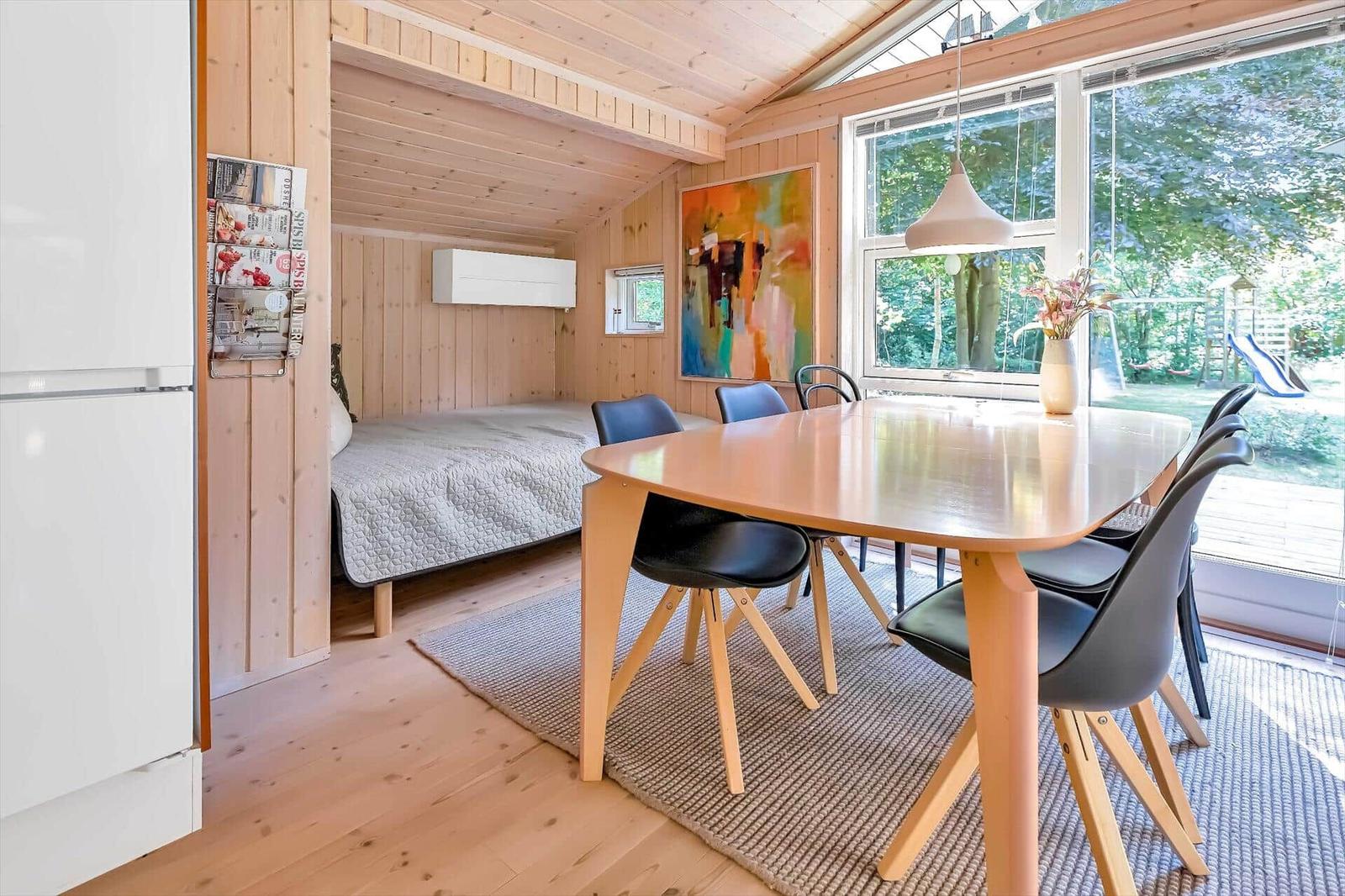 Essbereich mit Tisch und Stühlen, Schlafbereich im Hintergrund, große Fenster mit Blick auf Garten.