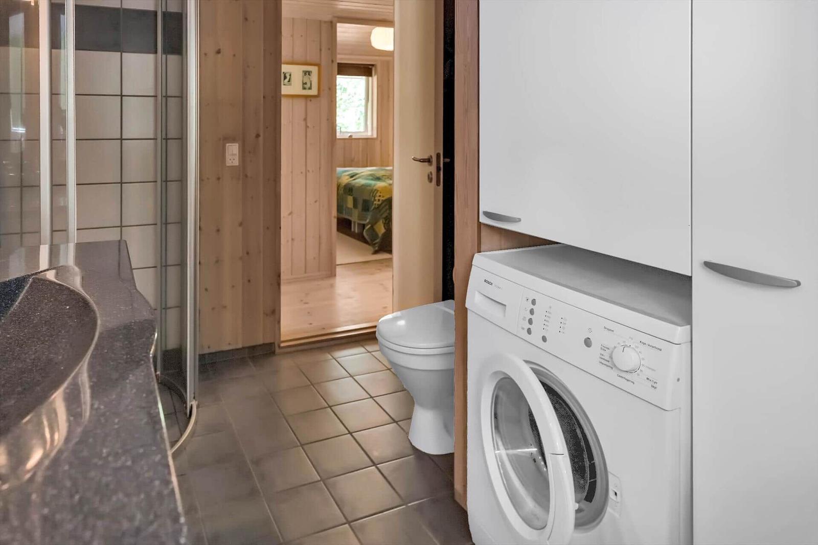 Badezimmer mit Waschmaschine, Toilette und Dusche. Blick in Schlafzimmer mit Bett.