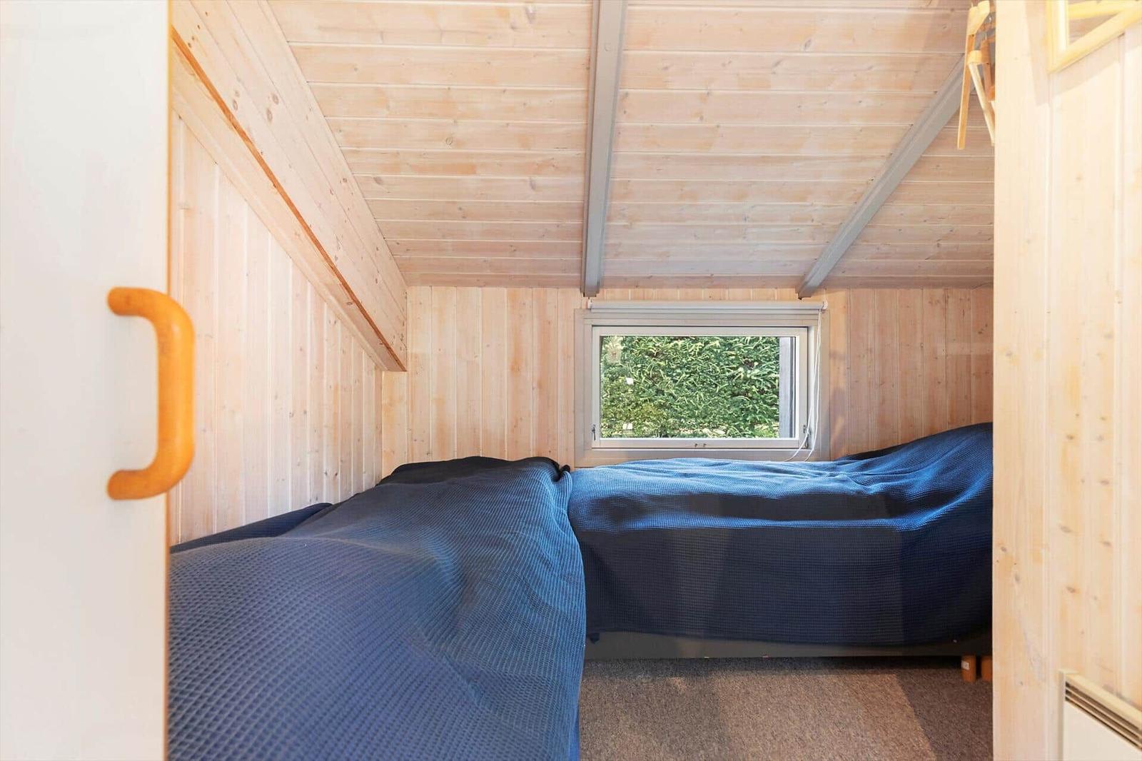 Schlafzimmer mit Holzwand und blauer Bettdecke. Fenster zeigt grüne Vegetation.