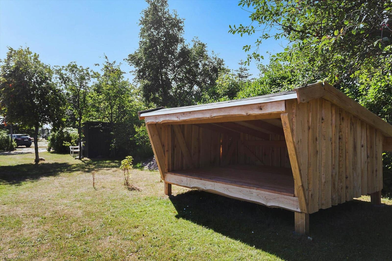 Holzüberdachung mit Sitzfläche im Garten. Umgeben von Bäumen und Gras.