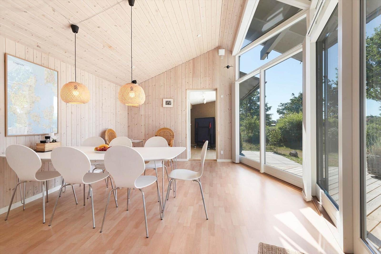 Esszimmer mit weißem Tisch, Stühlen und Holzwand. Große Fenster mit Blick auf Garten.