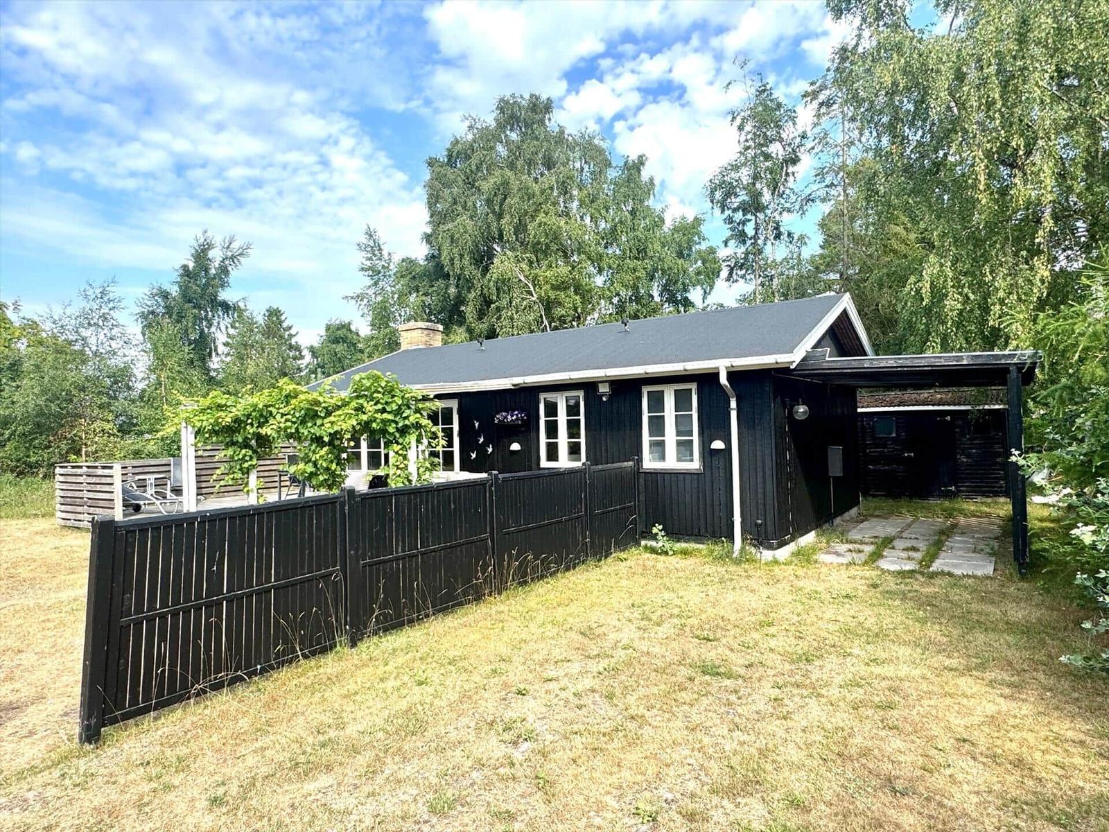 Black wooden house with white window frames and garden fence. Surrounded by trees and grass.
