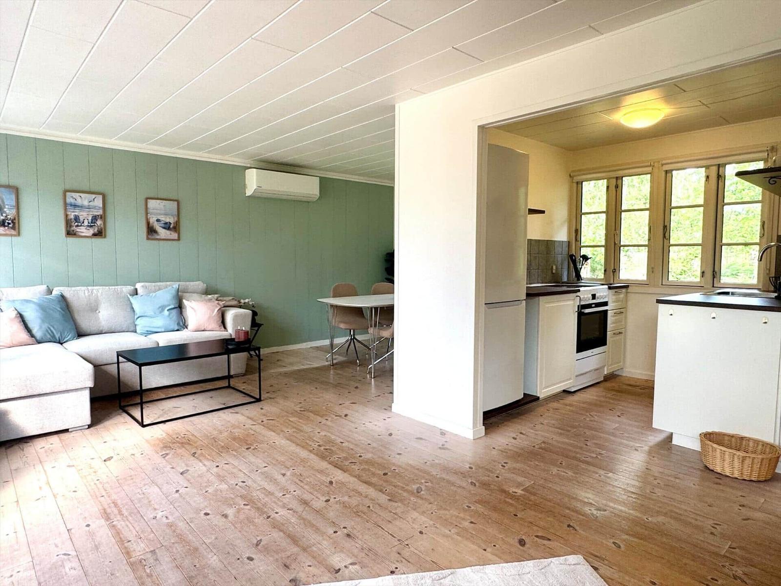 Living area with sofa, table, and kitchen. Wooden floor, green walls, windows with view of greenery.