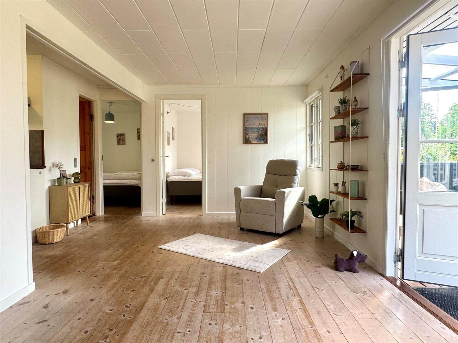 Living area with armchair, wooden floor, and view into bedrooms. Shelving and window visible.