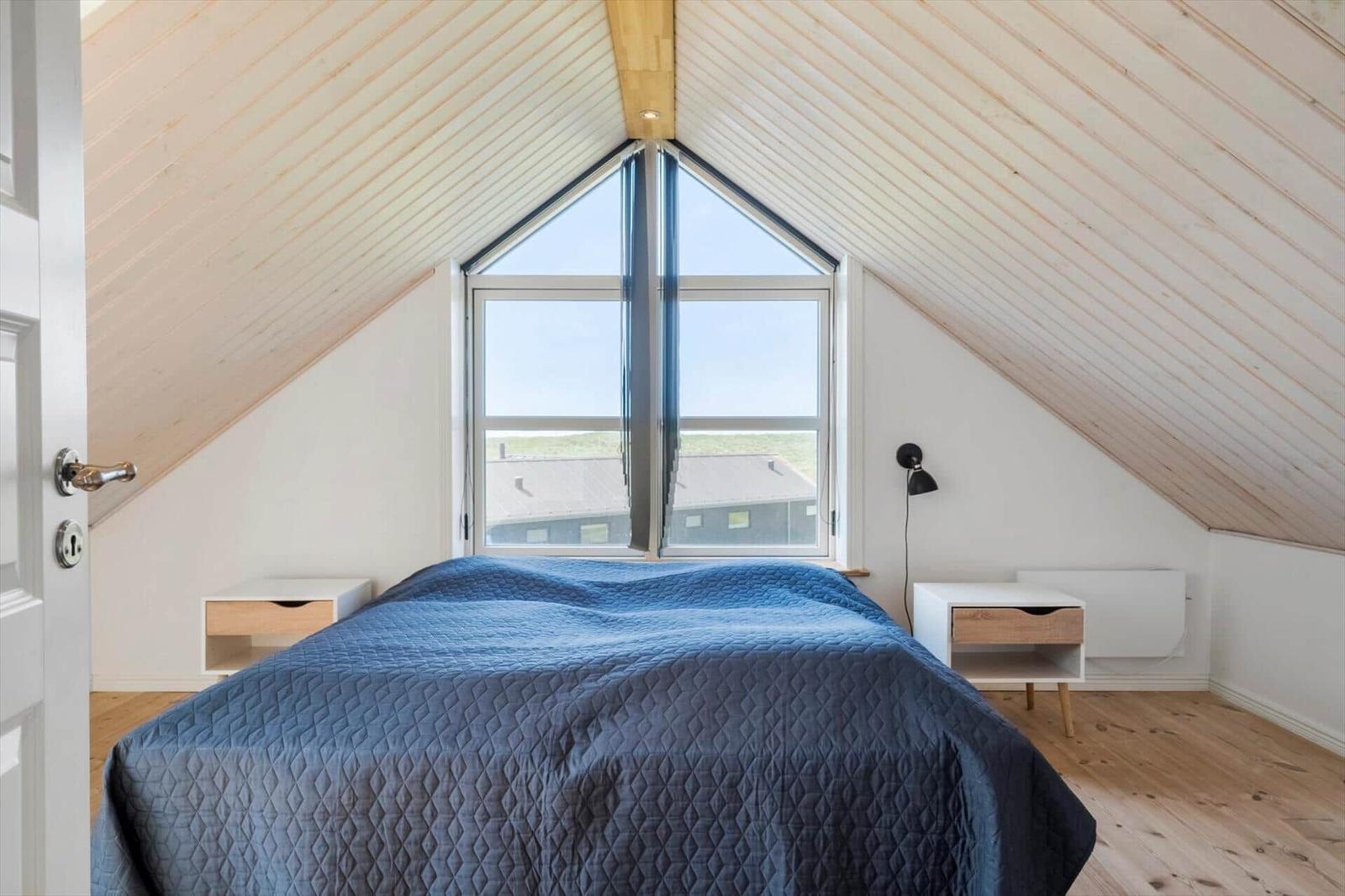 Schlafzimmer mit Dachschrägen, großem Fenster und blauem Bettbezug.
