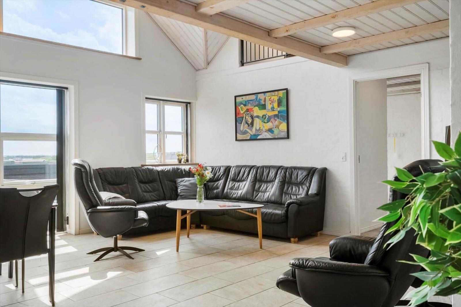 Wohnzimmer mit schwarzer Ledercouch, Holztisch und großen Fenstern. Deckenbalken und Wandbild sind sichtbar.