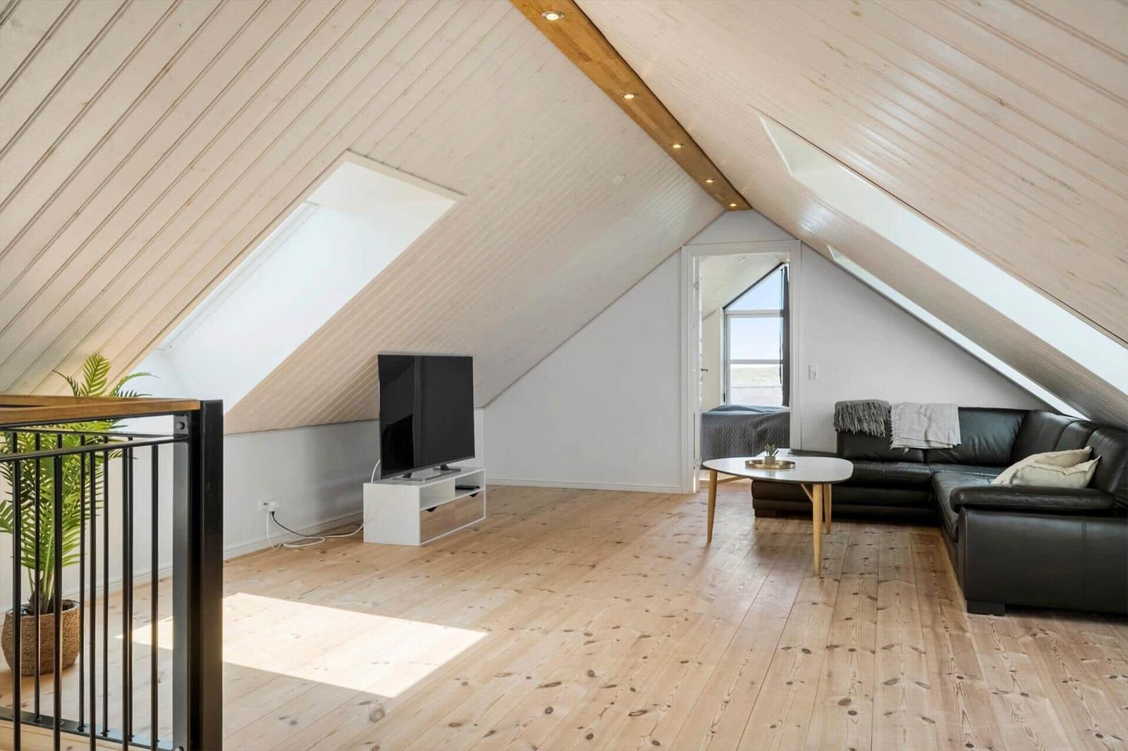 Wohnraum mit Holzboden, schwarzem Sofa und Fernseher unter schrägem Dach