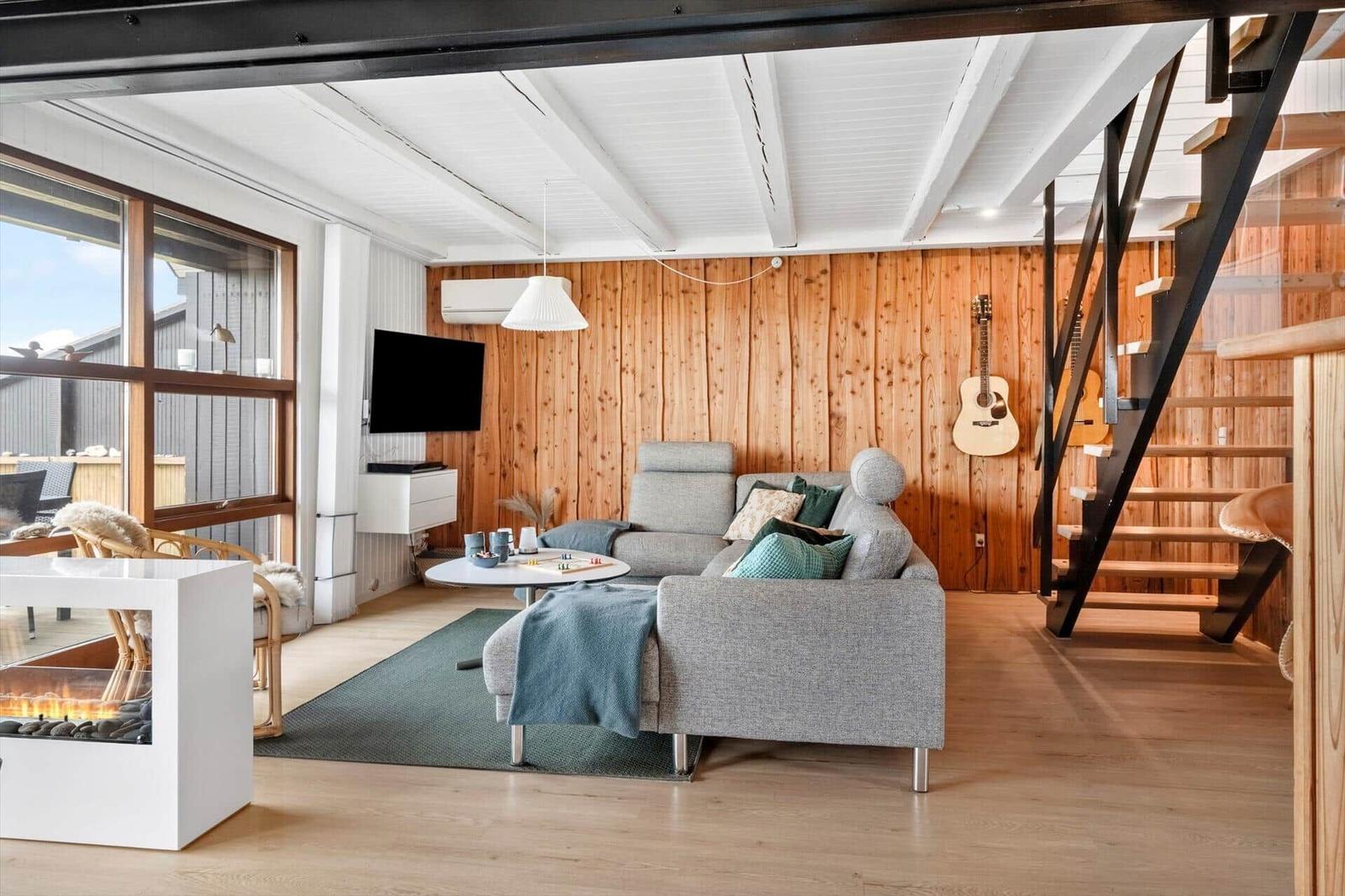 Wohnzimmer mit Sofa, Fernseher und Holzwand. Treppenhaus und Kamin sichtbar.