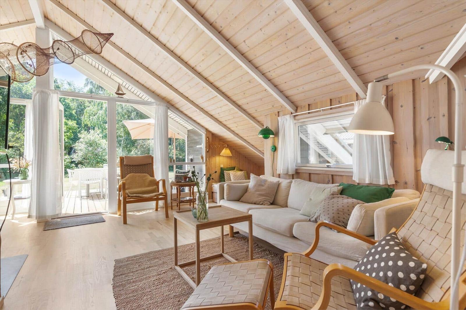 Wohnraum mit Sofa, Sesseln und Holzdecke. Durchgang zur Terrasse mit Gartenblick.