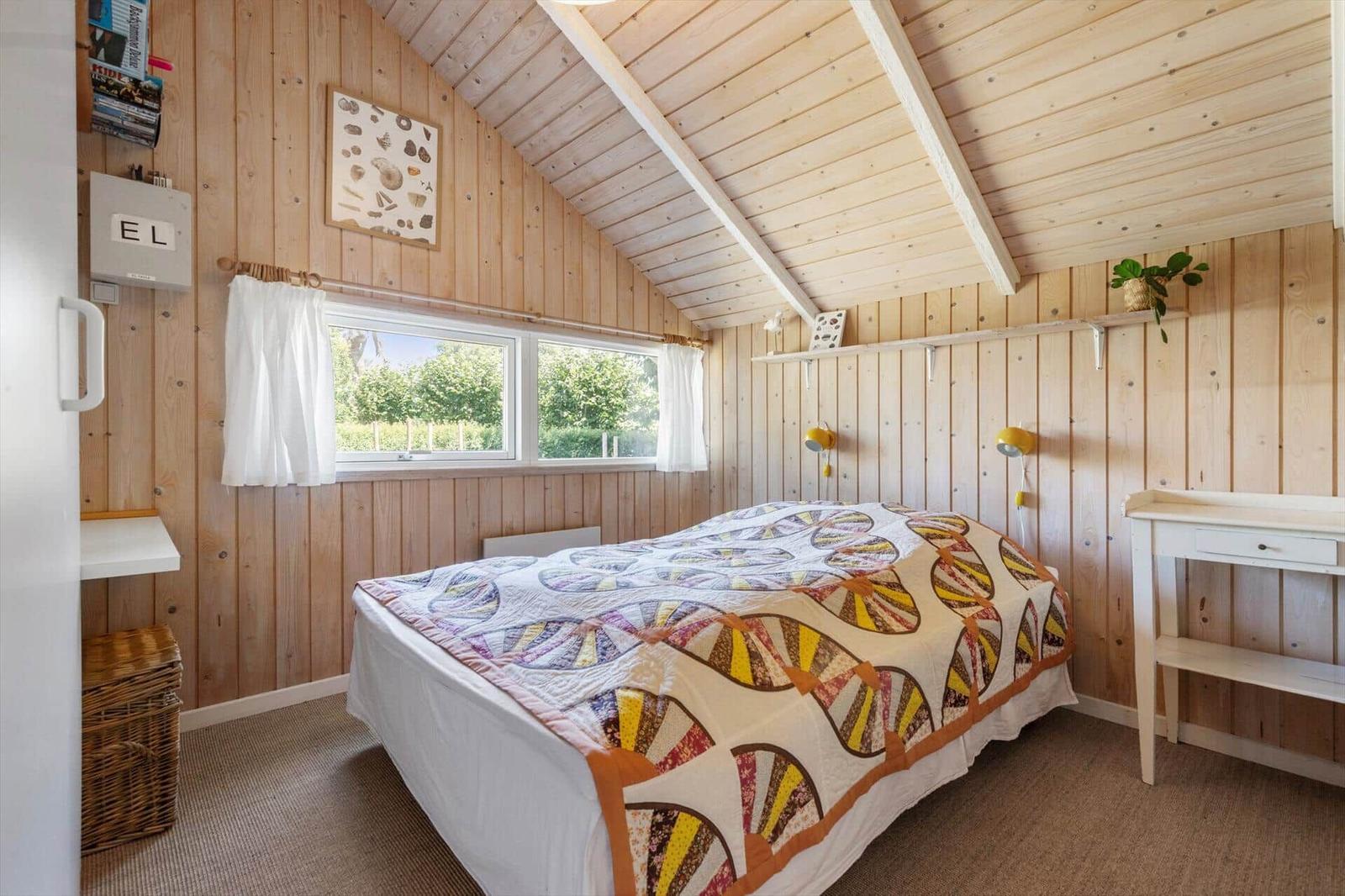 Schlafzimmer mit Holzwand, Bett mit buntem Decken, Fenster mit Blick auf Grün, Schreibtisch und Pflanze.