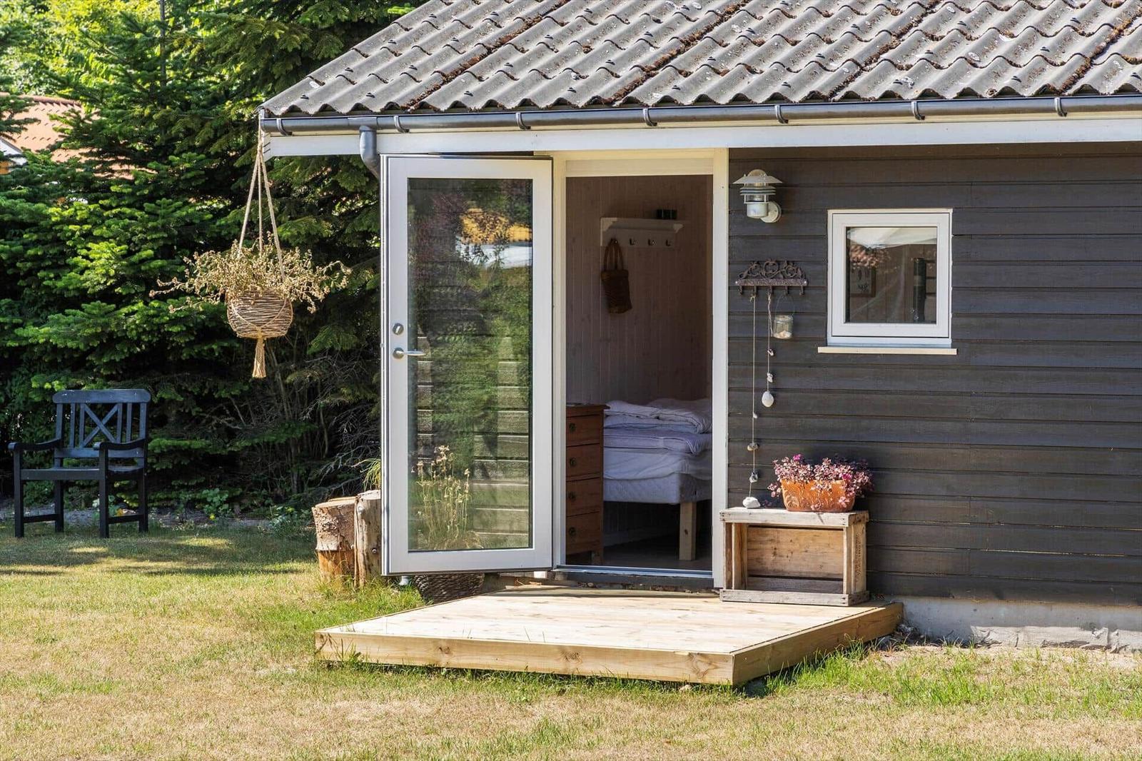 Haus mit offener Tür, darin Bett und Schrank. Außen Holzterrasse, Stuhl und Blumenkästen.