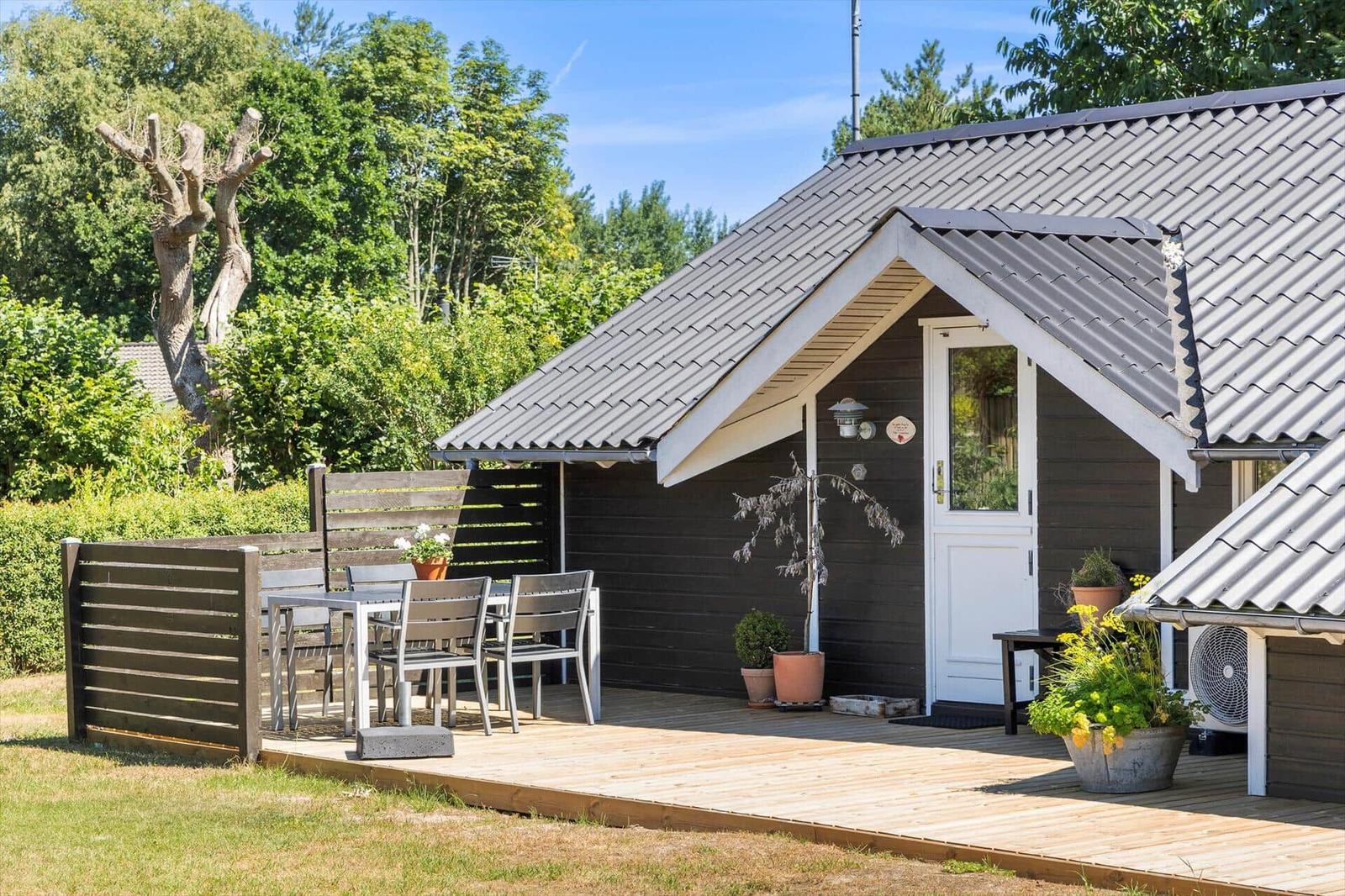 Hütte mit Holzterrasse, Tisch und Stühlen, Pflanzen und weißer Tür.
