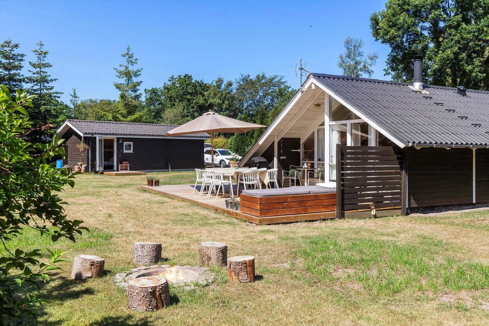 Haus mit Terrasse, Grill und Außentisch unter Sonnenschutz. Feuerstelle mit Baumstumpf-Sitzbänken.