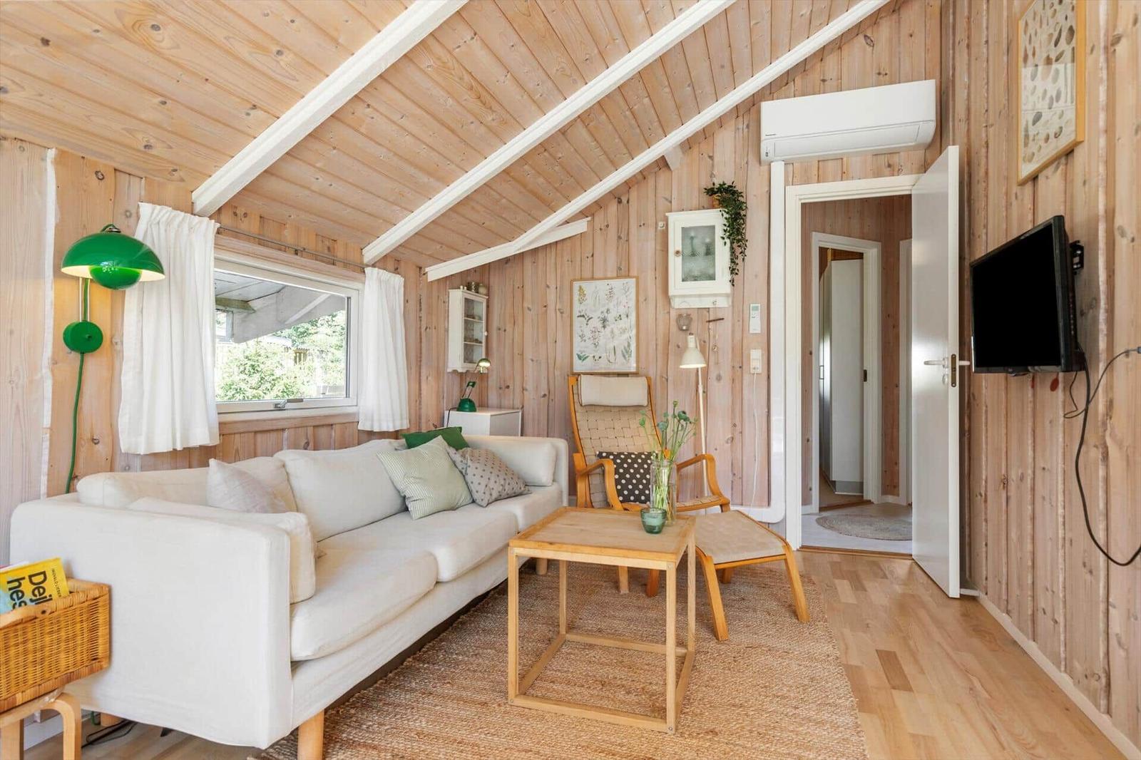 Wohnzimmer mit Sofa, Tisch und Fernseher an Holzwand. Fenster mit weißen Vorhängen.