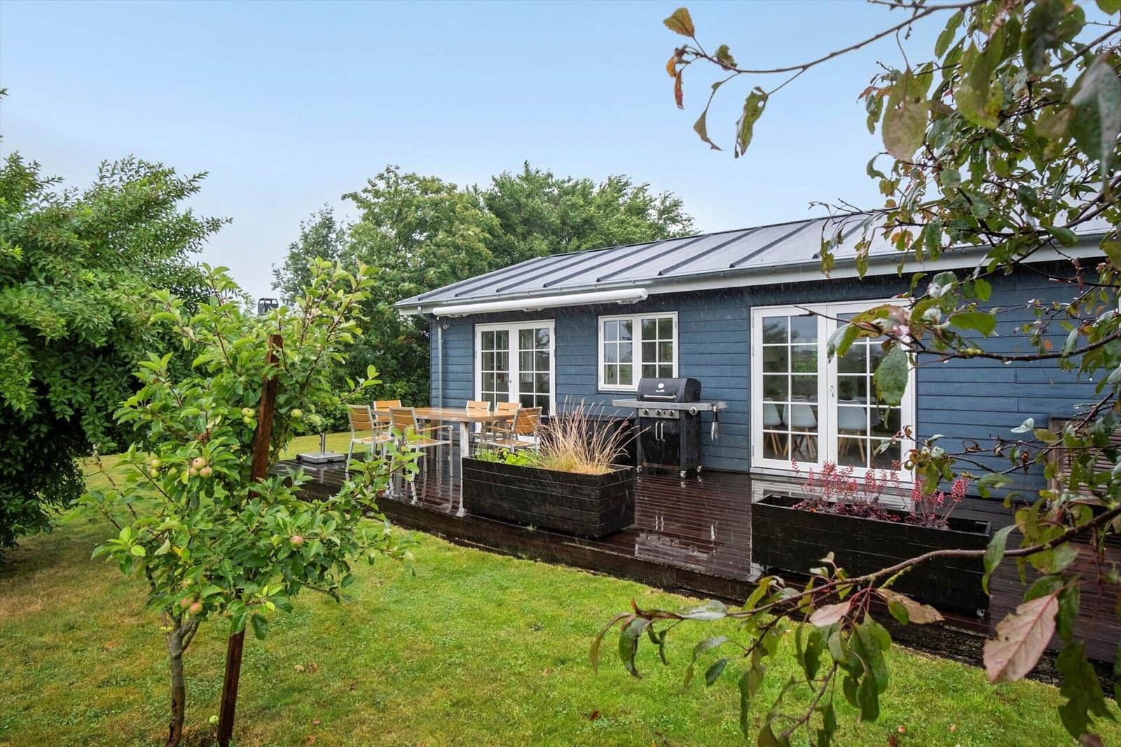 Blue wooden house with terrace, grill, and garden. Trees and grass surround the house.