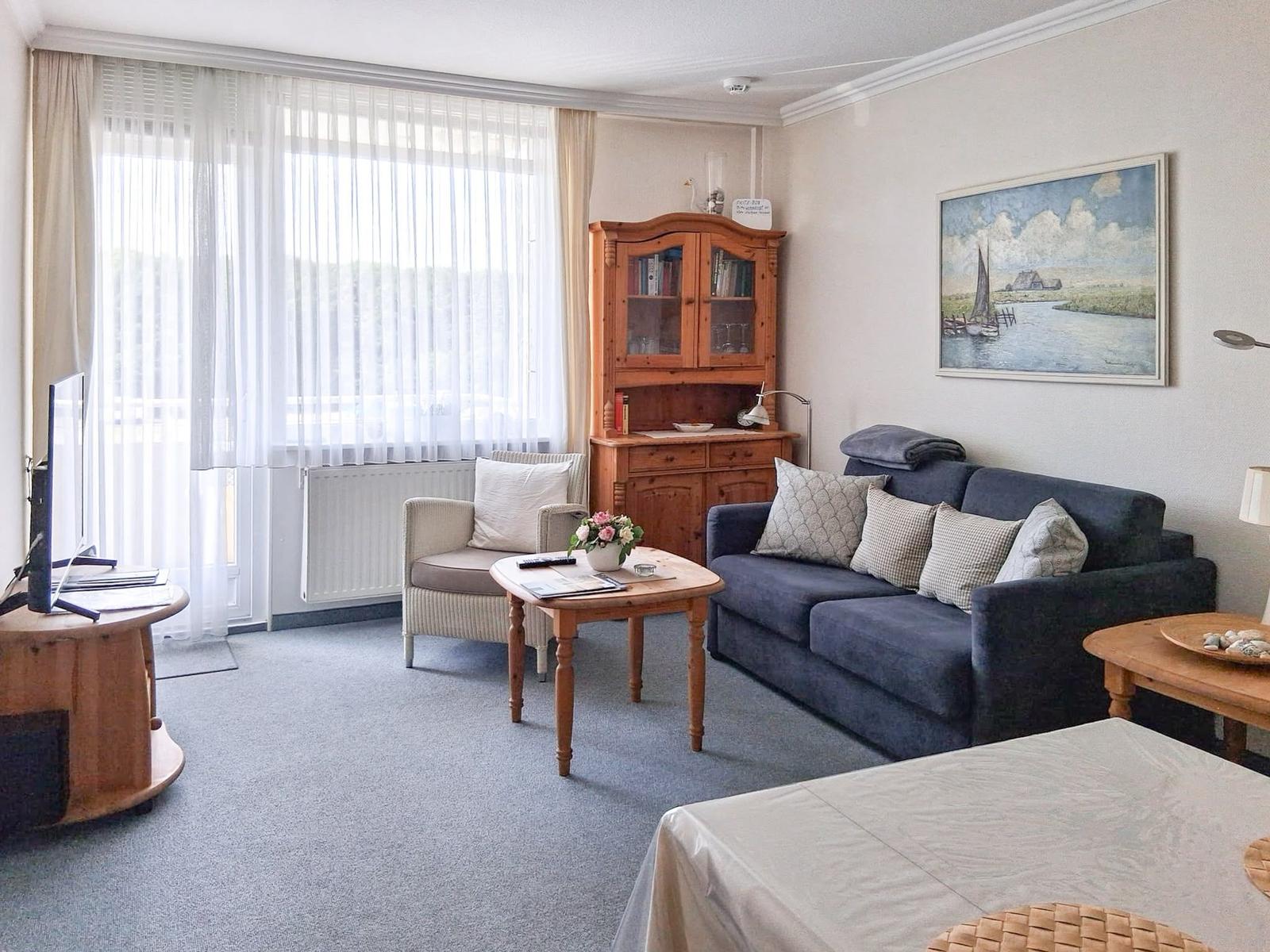 Wohnzimmer mit Sofa, Sessel, Tisch und Fernseher. Fenster mit Vorhängen und Wandbild.