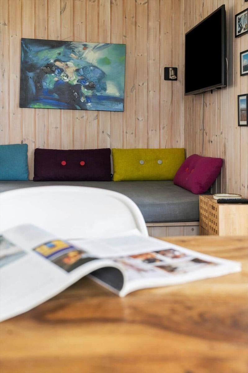 Ein Raum mit Holzwand, Sofa mit bunten Kissen und Wandbild. Auf Tisch liegt eine Zeitschrift.