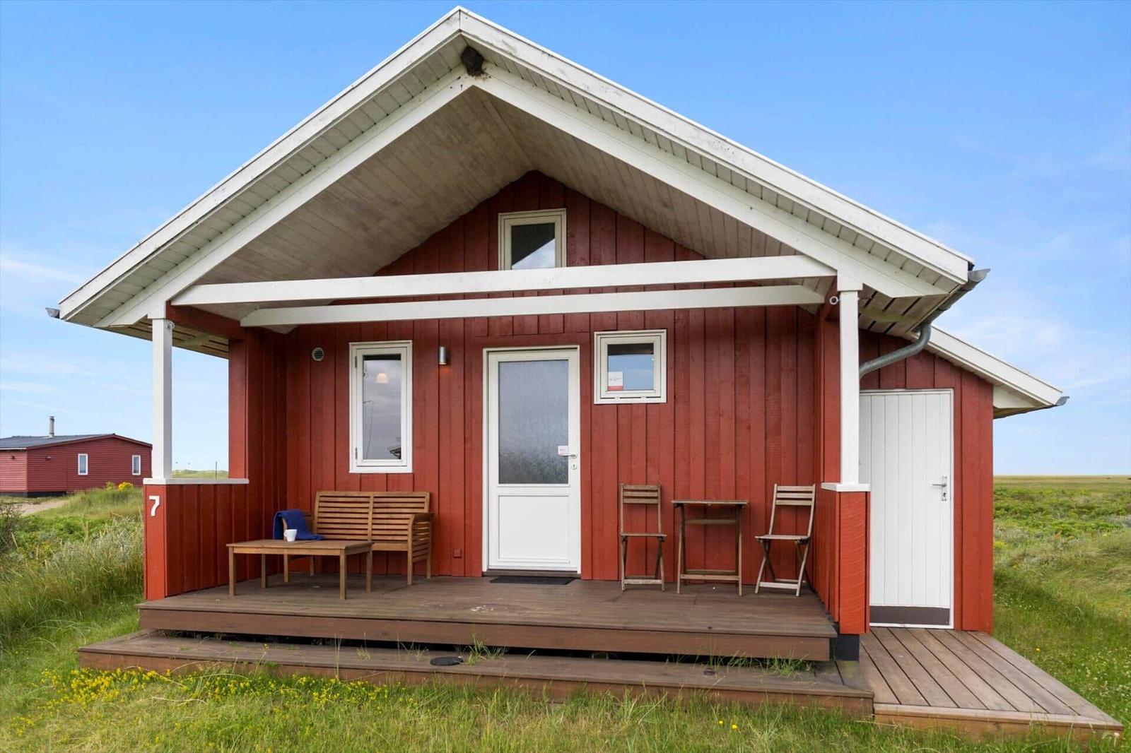 Rotes Holzhaus mit Terrasse, Tisch, Stühlen und Fenstern. Grasland im Hintergrund.