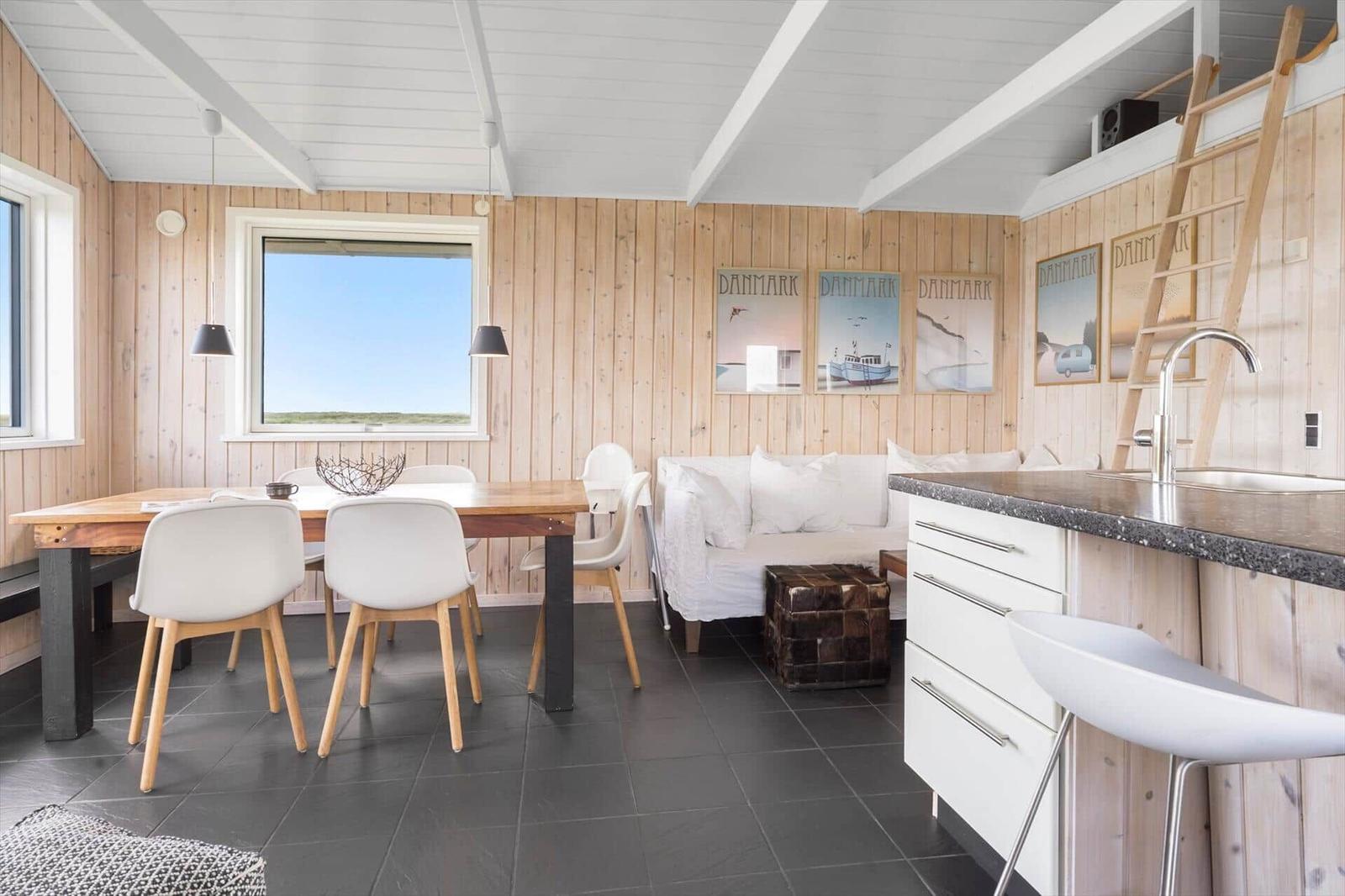 Wohnraum mit Essbereich, Sofa und Küchenzeile. Holzwände, Fenster und Tisch.