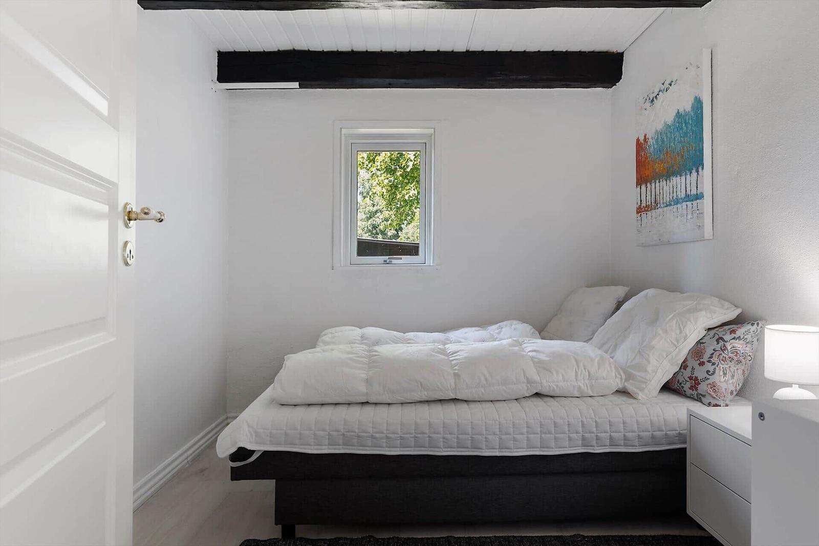 Schlafzimmer mit Bett, Nachttisch und Fenster. Deckenbalken und Wandbild sichtbar.