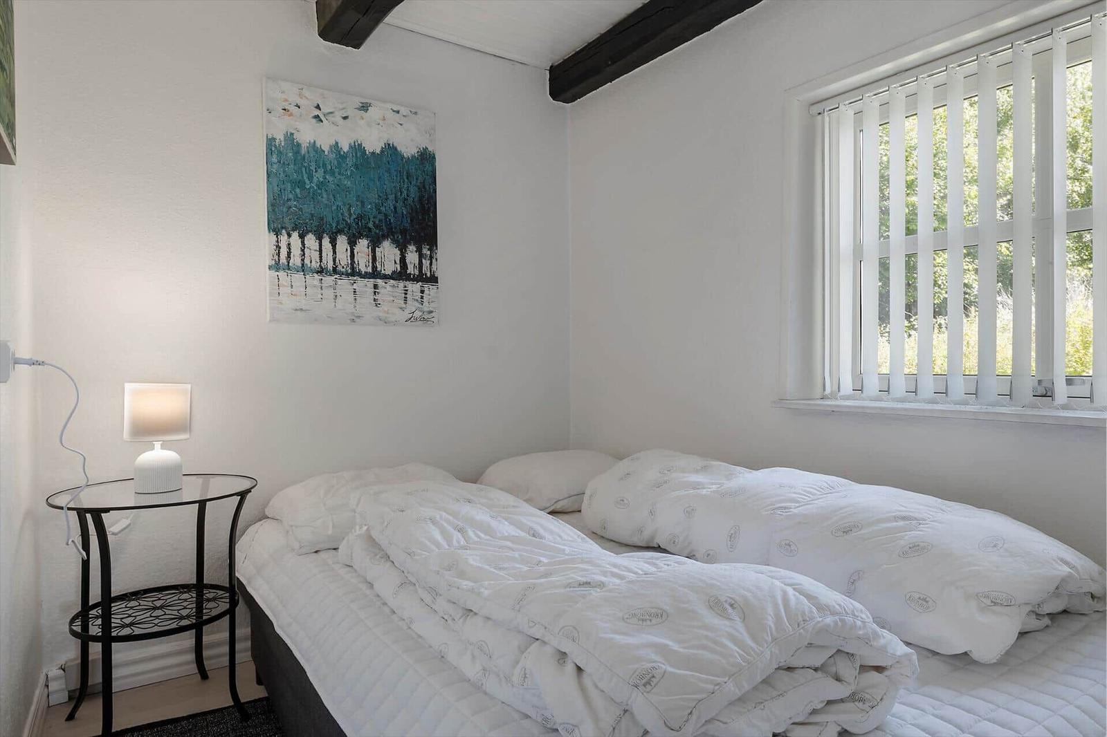 Schlafzimmer mit Bett, Nachttisch und Fenster mit Jalousie.