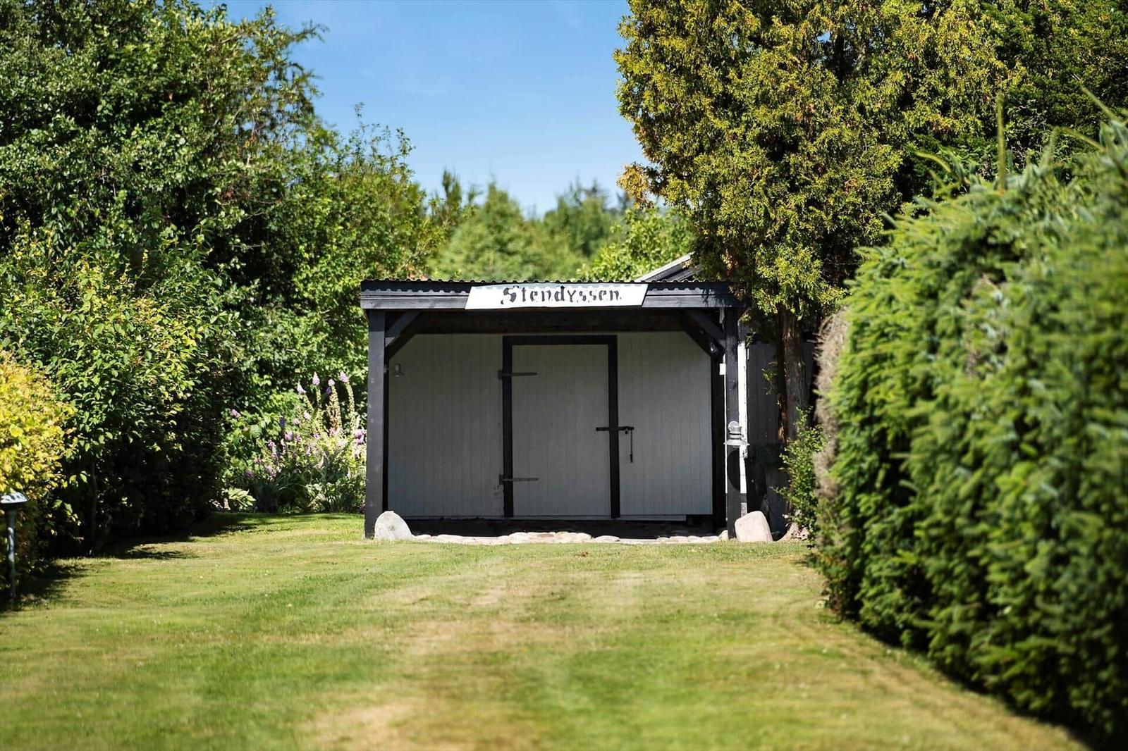 Ein weißer Schuppen mit schwarzen Rahmen und Schild Stendyssen steht im Gras zwischen Bäumen.