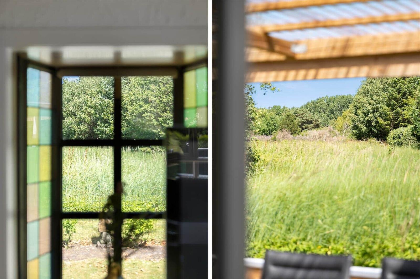Durch Fensterblick auf grüne Wiese und Bäume. Links: farbiges Glasfenster. Rechts: Sicht unter Dachkonstruktion.