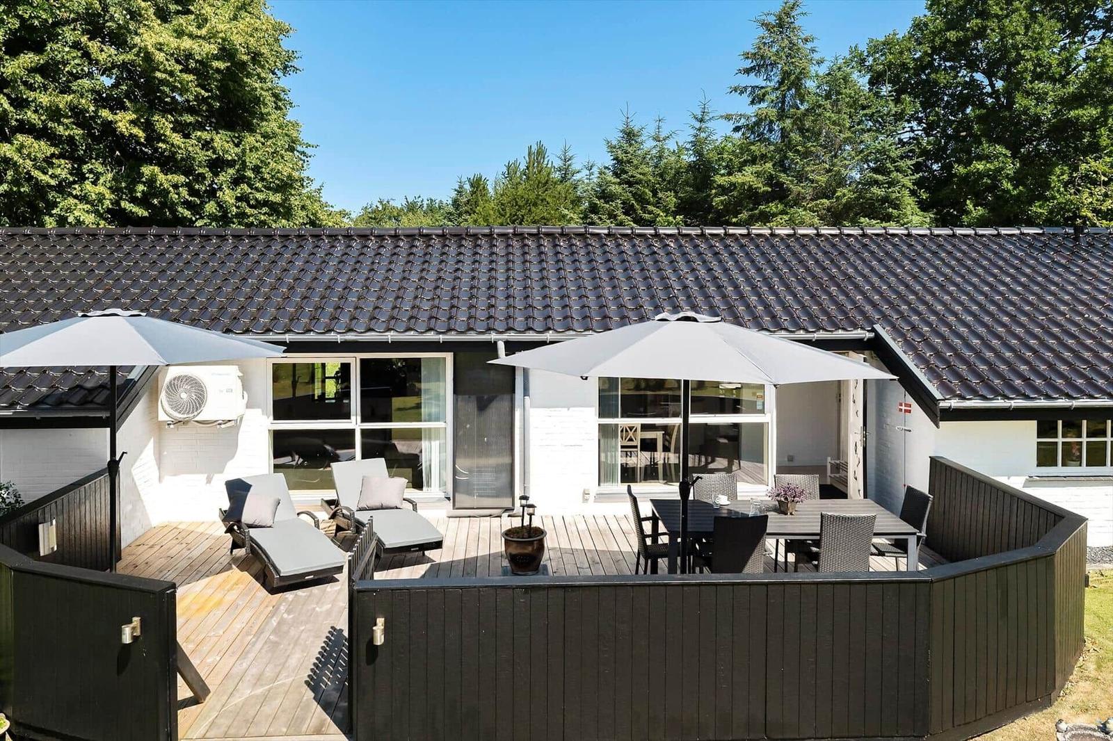 Terrasse mit Liegen, Tisch und Sonnenschirmen. Haus mit dunklem Dach und weißen Wänden.