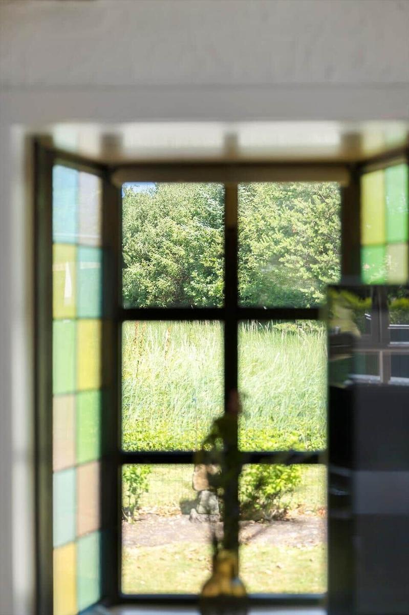 Durch ein Fenster mit bunten Glasflächen blickt man auf einen grünen Garten mit Bäumen.