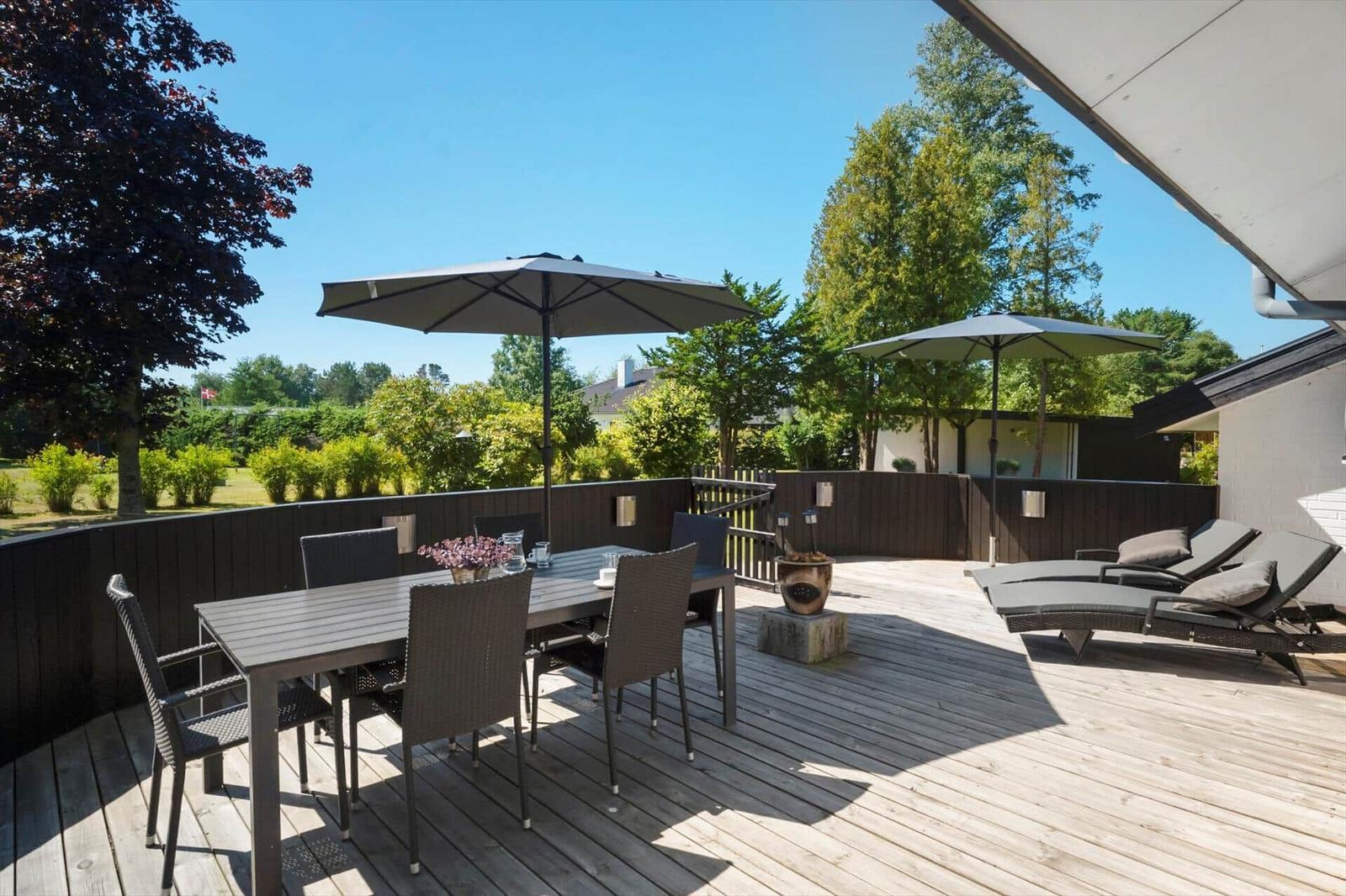 Terrasse mit Tisch, Stühlen, Sonnenschirmen und Liegen. Holzdecke und grüne Umgebung.