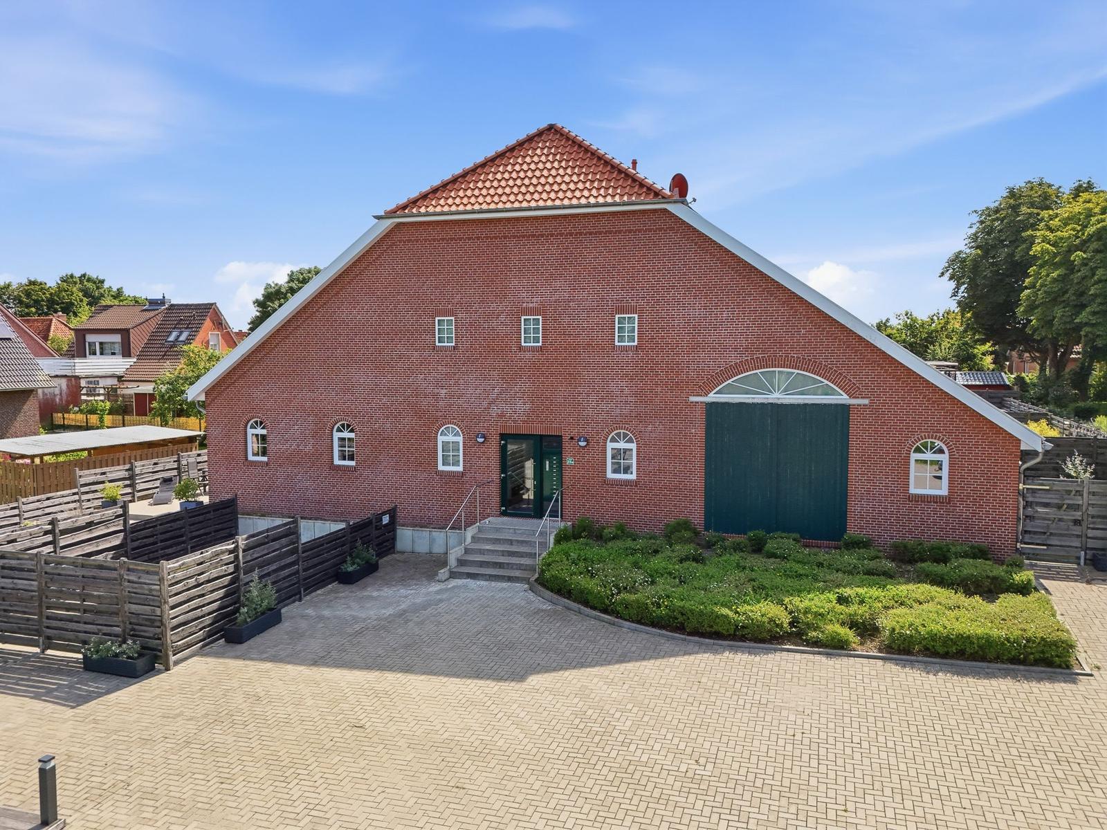 Rotes Backsteinhaus mit grüner Tür und gepflastertem Hof. Umgeben von Grünflächen und Holzzaun.