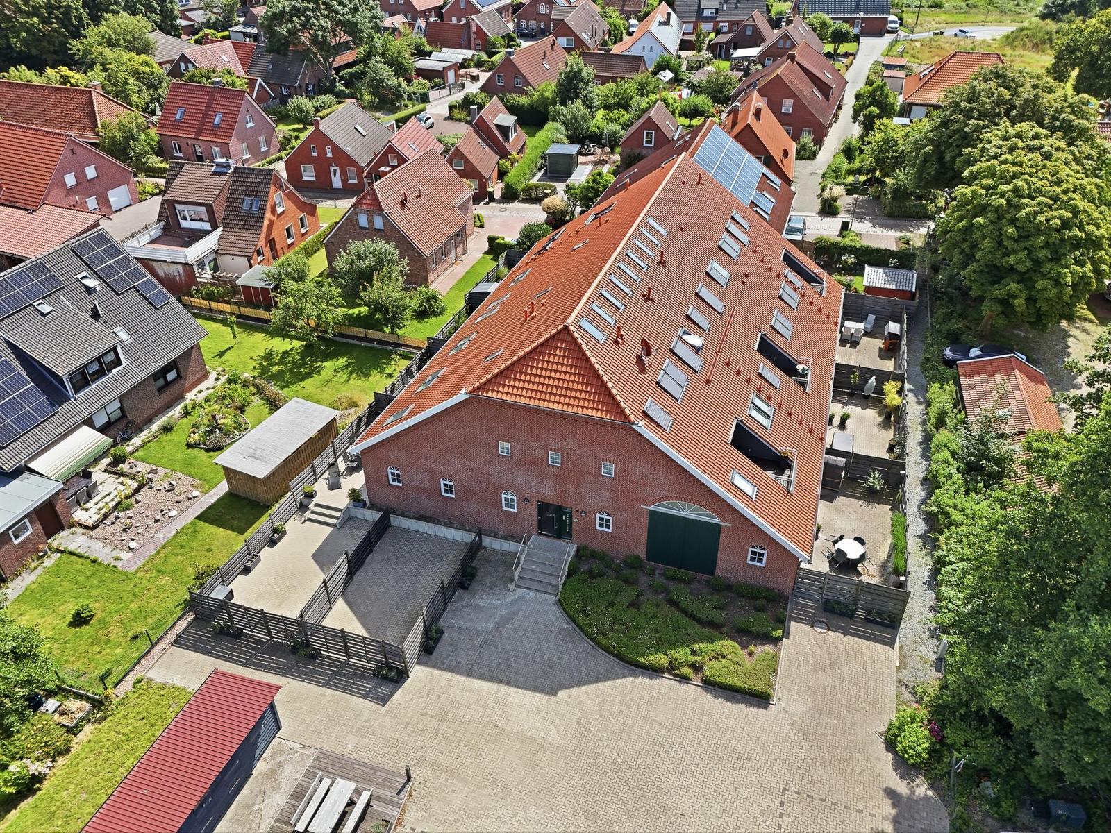 Roter Ziegelbau mit Solaranlage und gepflastertem Hof. Umgeben von Grünflächen und Nachbarhäusern.