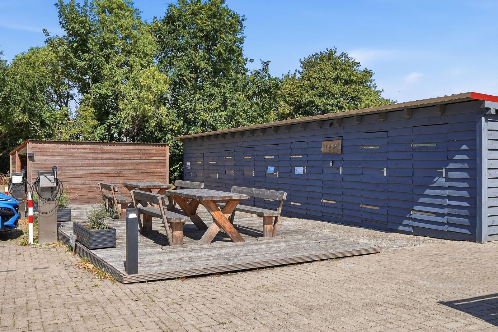 Holzterrasse mit Tisch und Bänken vor blauem Schuppen. Hintergrund: Bäume und klarer Himmel.