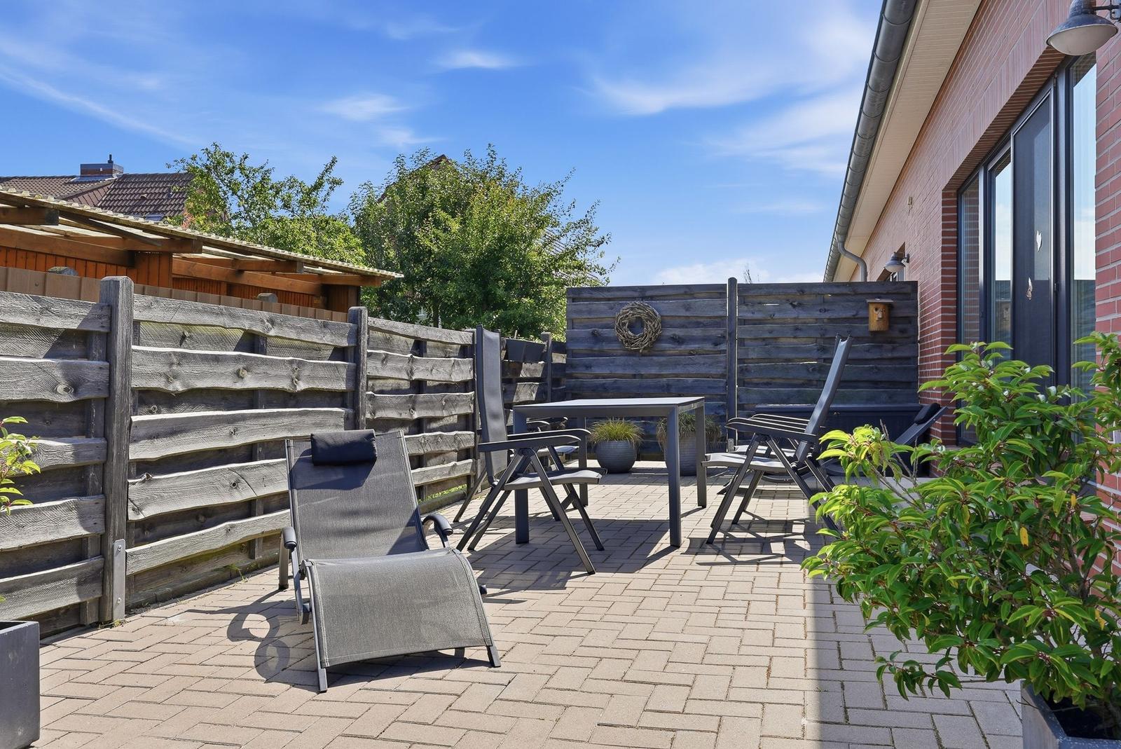 Terrasse mit Sitzgelegenheiten, Holzumrandung und Pflanzen. Hintergrund: Haus und Bäume.