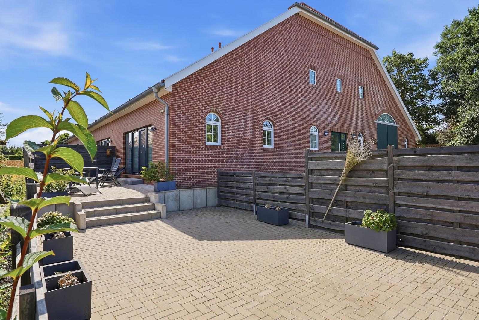 Roter Backsteinbau mit Terrasse und Holzzaun. Pflanzkübel und Sitzgelegenheiten.