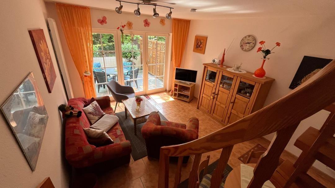 Wohnzimmer mit Sofa, Sessel und TV. Durchgang zur Terrasse mit Tisch und Stühlen.