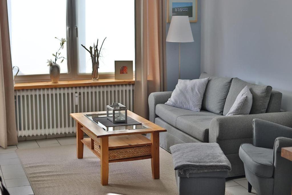 Ein Wohnzimmer mit grauem Sofa, Holz-Tisch und Fenster mit Blumen.