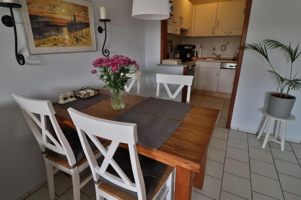 Essbereich mit Holztisch, weißen Stühlen und Blumen. Küche im Hintergrund. Pflanze auf Ständer.