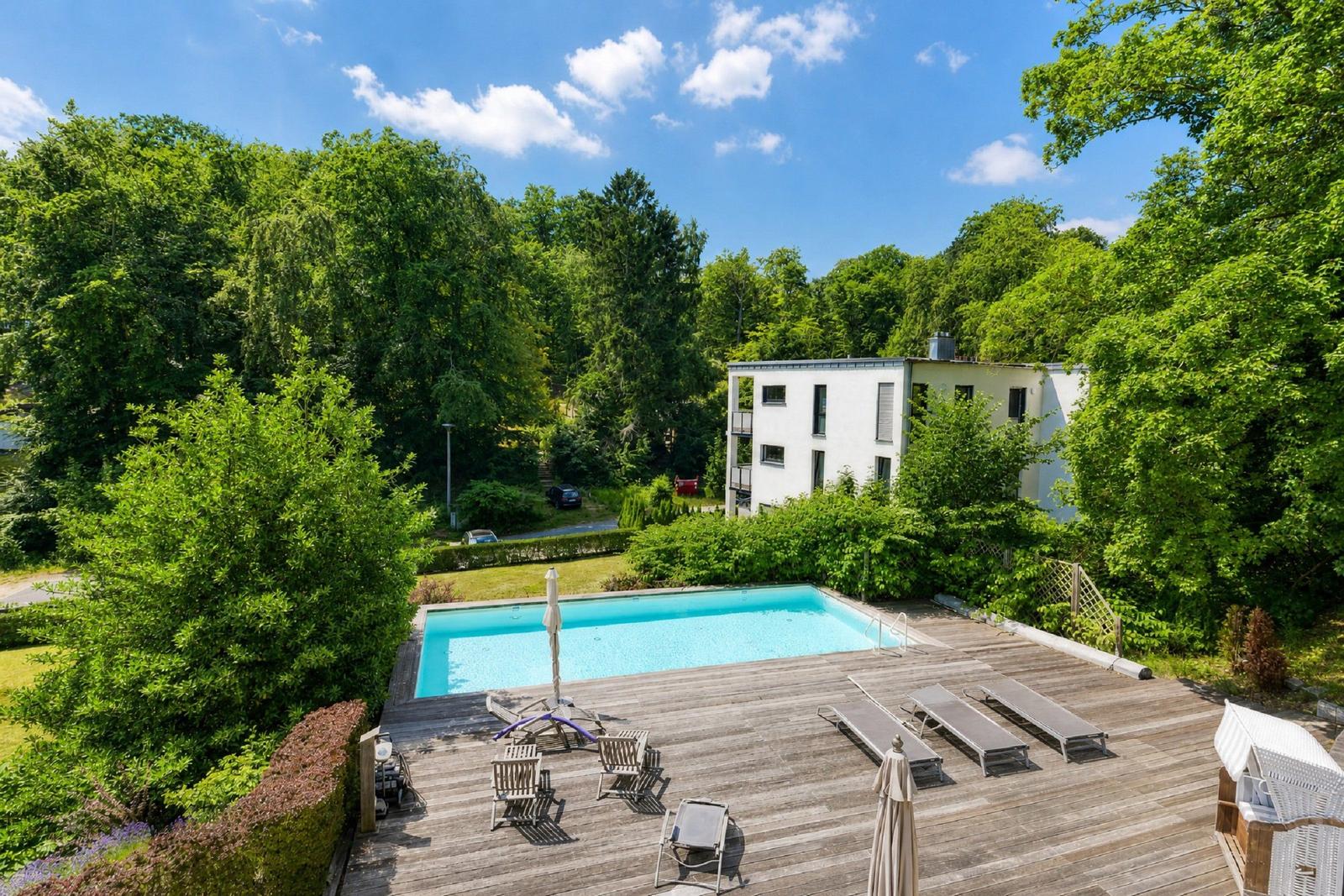 Pool mit Sonnenliegen und Holzterrasse umgeben von grüner Vegetation und einem weißen Gebäude.