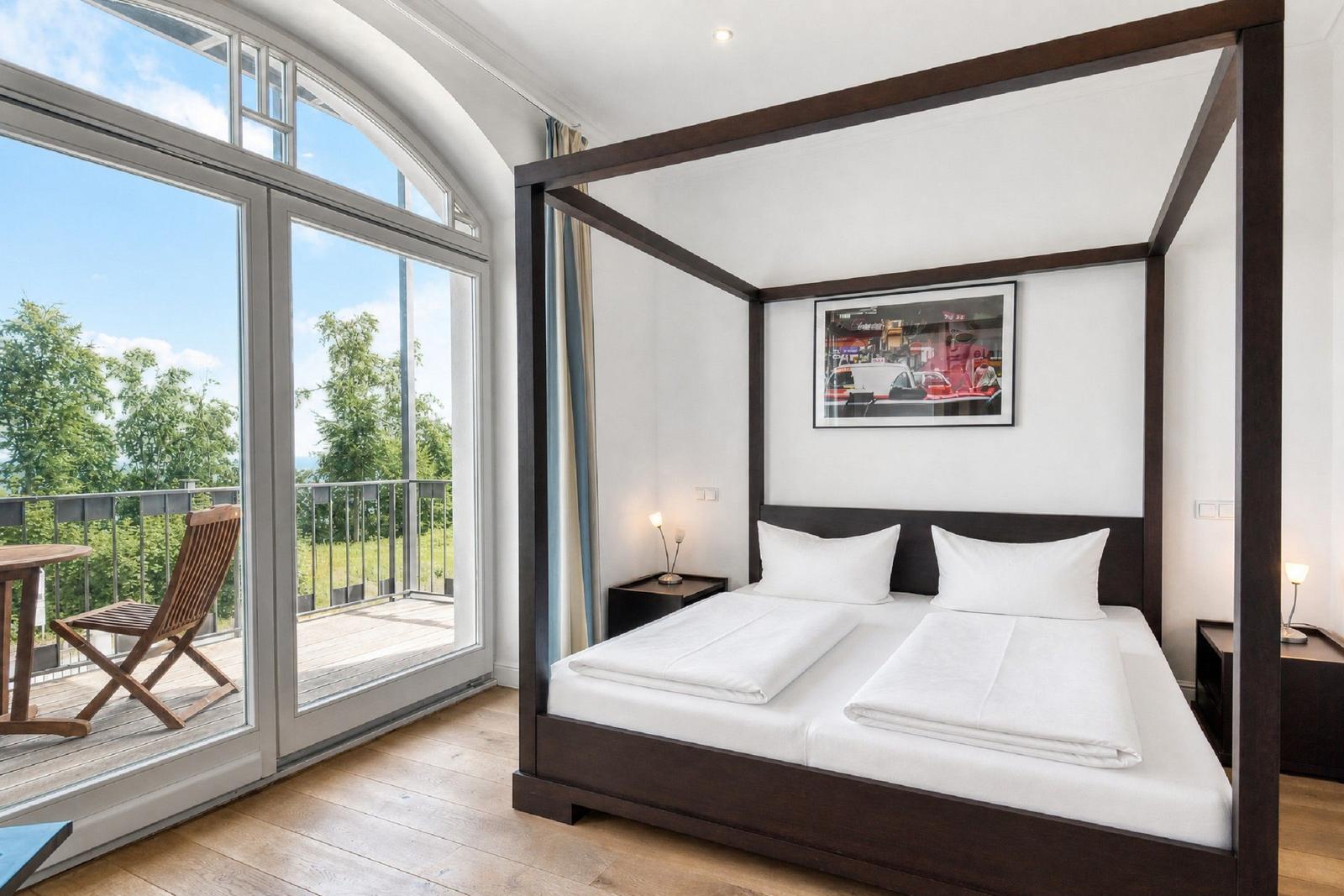 Doppelbett mit Balkonblick. Holzboden und gerahmtes Bild an der Wand.