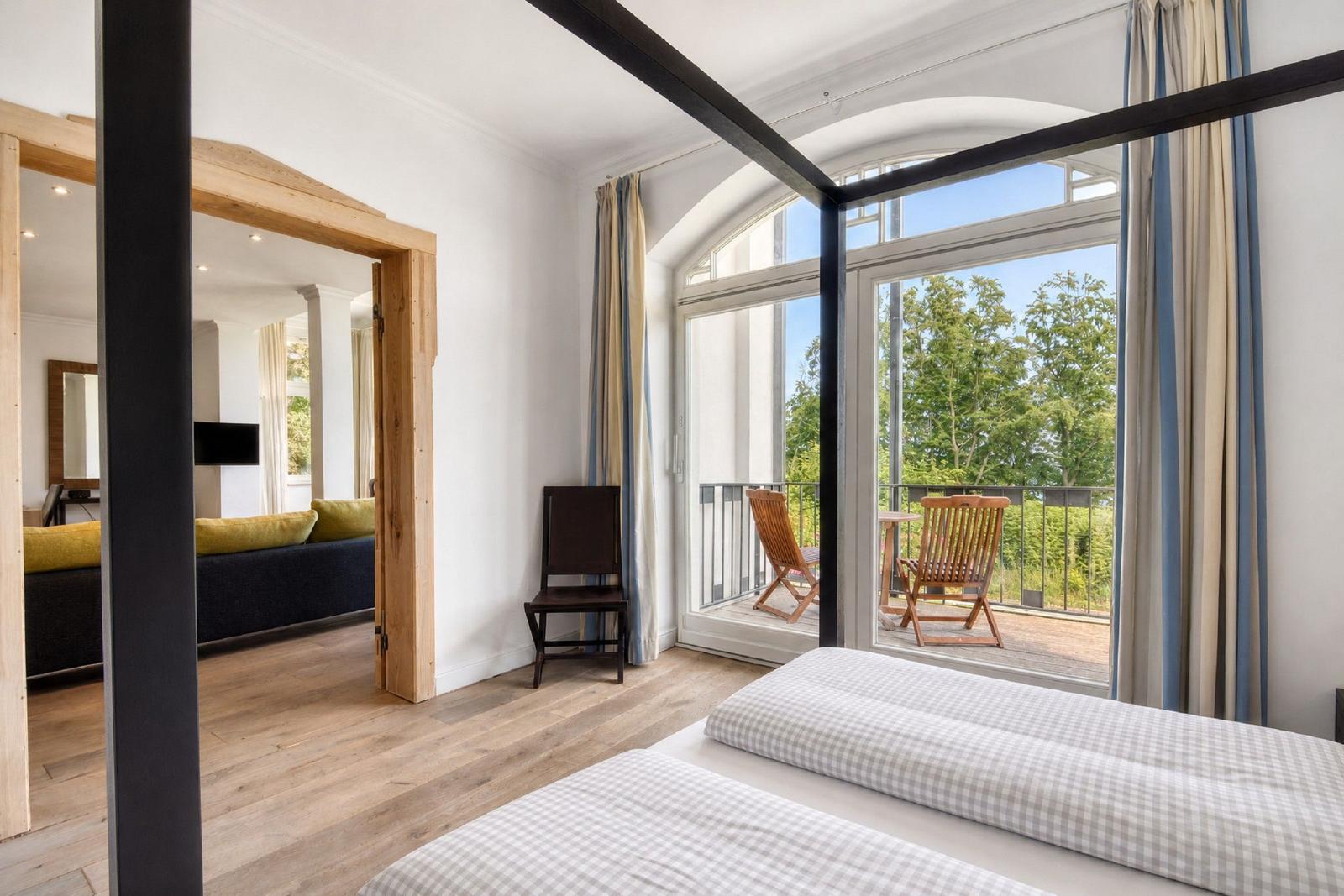 Schlafzimmer mit Balkon und Blick auf Bäume. Durchgang zur Wohnfläche mit Sofa.