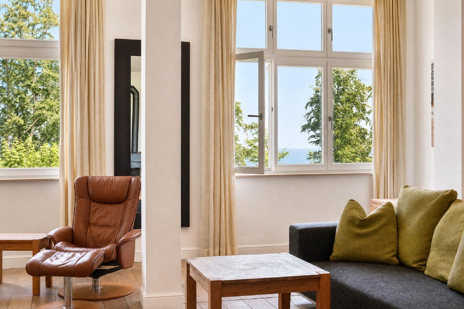 Wohnzimmer mit Sofa, Sessel und Blick auf Meer und Bäume durch Fenster.