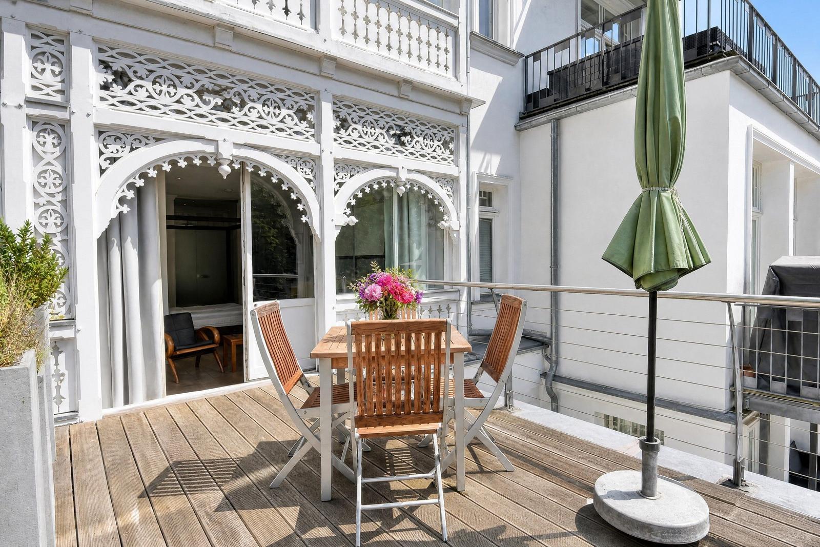 Terrasse mit Holztisch, Stühlen und grünem Sonnenschirm. Weiße Fassade mit kunstvollen Verzierungen.